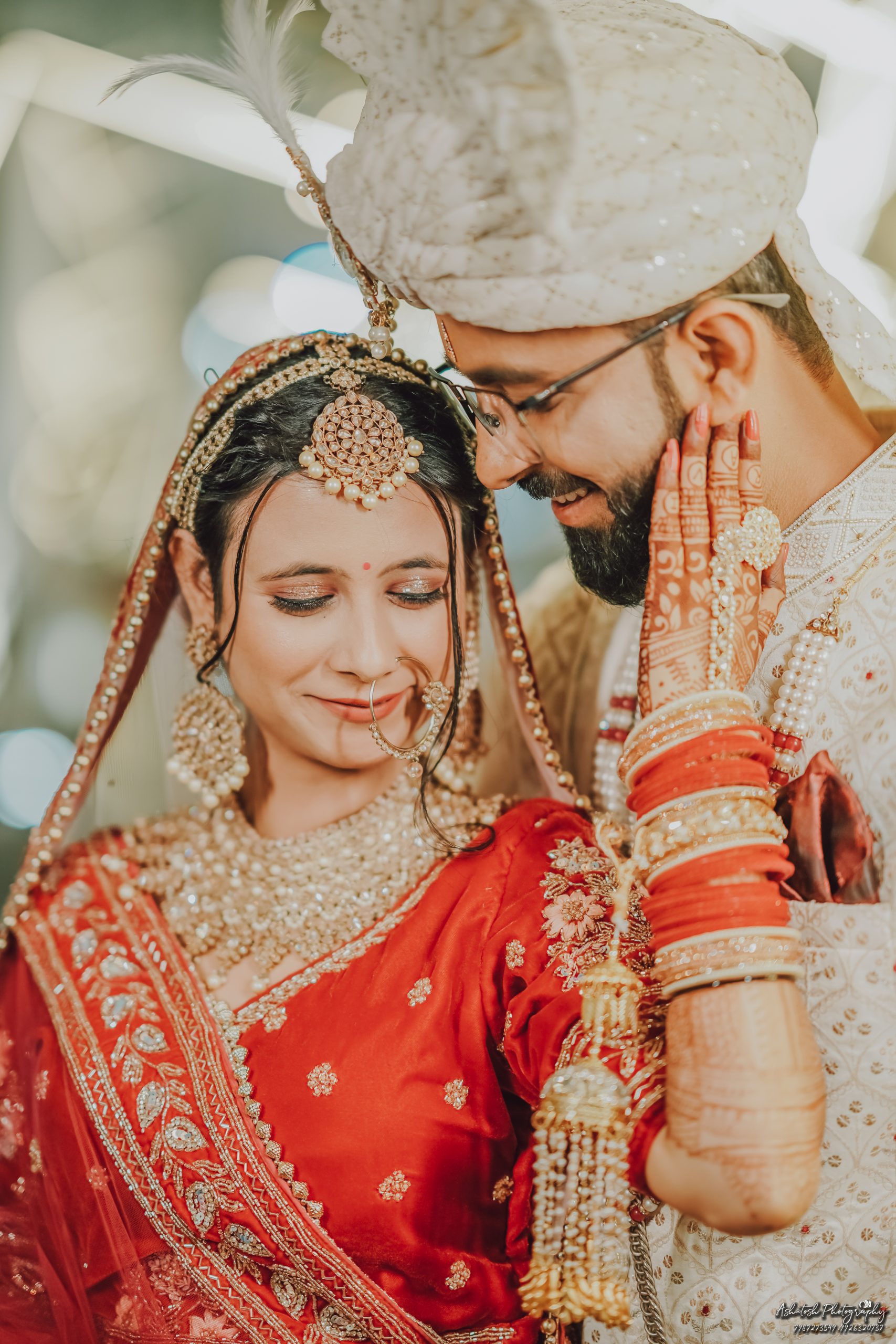 A close-up shot of a bride in a red traditional outfit adorned with intricately designed jewelry and embellishments, smiling softly while looking at her partner, who is wearing a cream-colored sherwani and a turban.