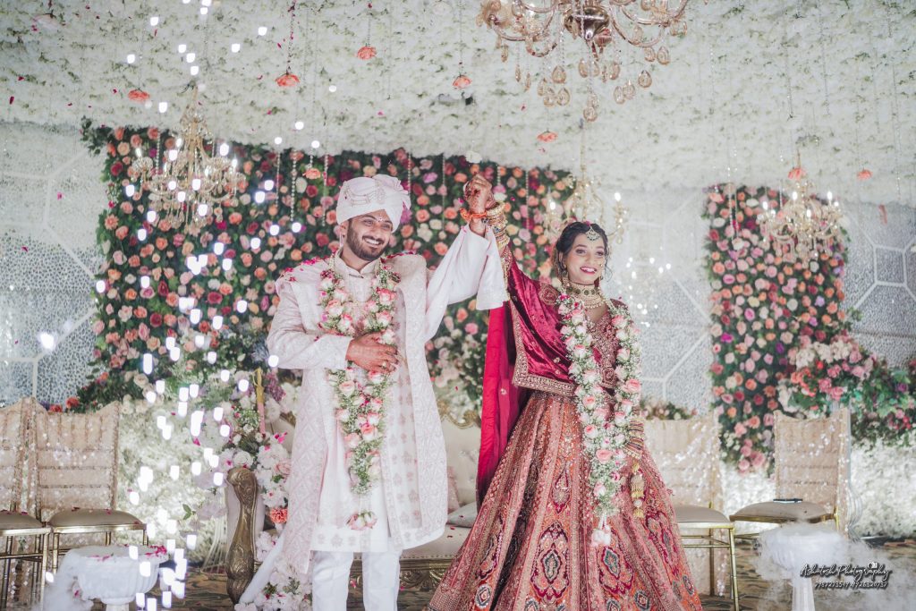 A joyous couple in traditional Indian wedding attire holds hands, surrounded by a lavish floral backdrop and elegant decor, celebrating their union.
