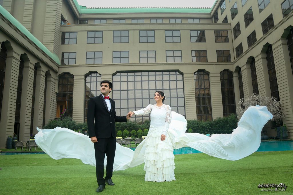 A bride and groom stand together on a lush green lawn, with the bride wearing a flowing white gown and the groom in a black suit with a red bowtie. Their attire billows gently in the breeze, set against the backdrop of an elegant building.