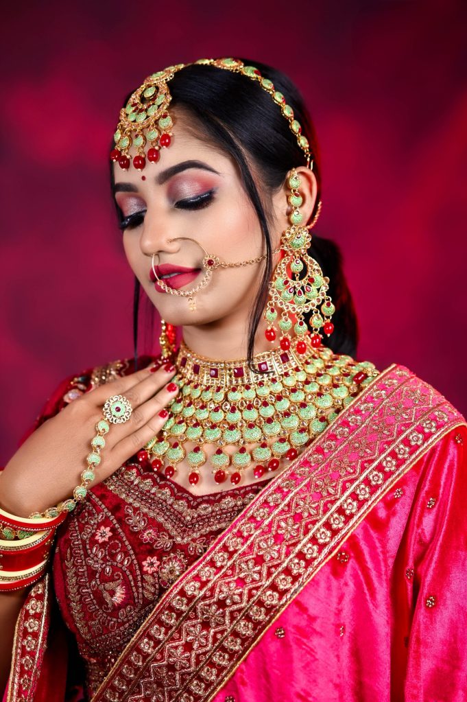 A woman wearing traditional Indian attire, adorned with elaborate jewelry, including a necklace and earrings, set against a vibrant red background.