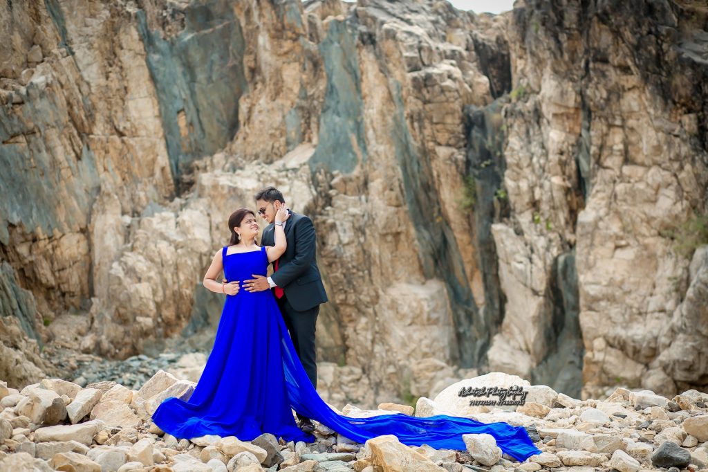 A couple embracing on rocky terrain, with the woman wearing a blue gown that flows around her, captured in a romantic moment.