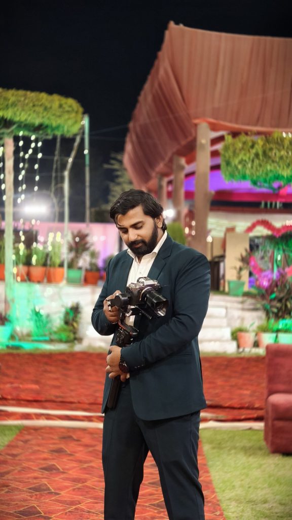A man in a dark suit holds a camera on a tripod, standing in a vibrant outdoor wedding setting with colorful lighting and decorations.