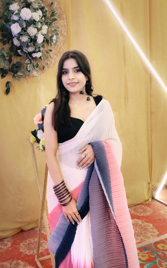 A woman elegantly dressed in a multi-colored pleated saree, adorned with bangles and rings, standing in front of a floral backdrop, exuding grace and confidence.