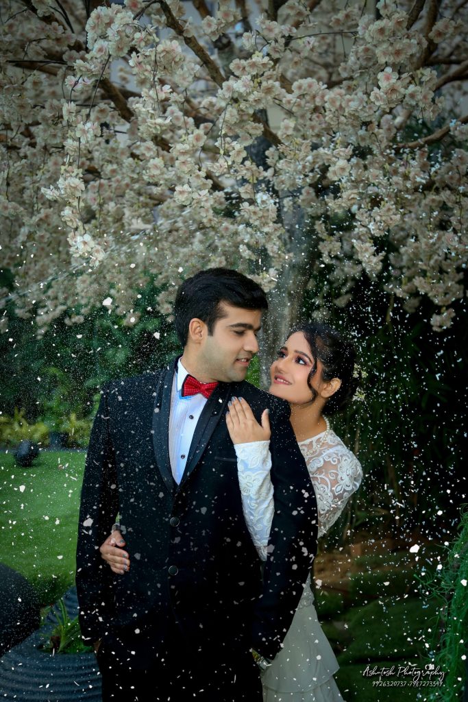 A couple standing close together under a flowering tree, surrounded by soft white petals falling like snow. The man is wearing a black suit with a bow tie, while the woman is dressed in an elegant white gown and looks up affectionately at him.
